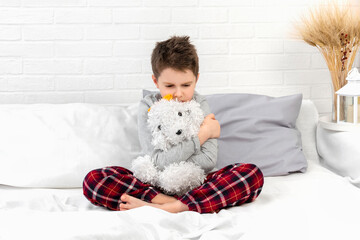 Obraz premium Angry boy in pajamas in bed. Child with a toy in bedroom at home in the morning. Sad schoolboy hugs teddy dog. Resentment, anger, annoyance, misunderstanding, bad mood, kids emotion, care, childhood