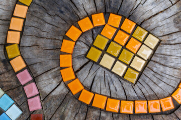 Mosaic colorful snake path. Abstract natural wooden background. Mosaic tiles on round cut down tree with cracks stump outside. DIY garden furniture, decorated by hand made element small tiles