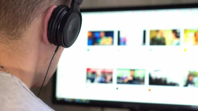 A young guy with headphones in the room looks at the computer monitor and surfs the Internet. Passes online training or work online.