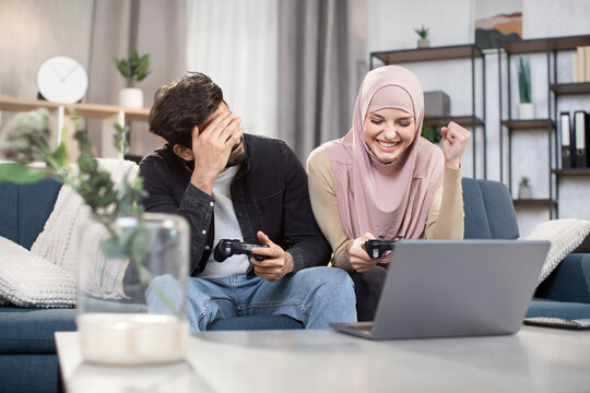 Excited funny multiehnic Muslim couple having fun together while playing video games at home. Laughing woman in hijab celebrating victory, while her friend is sad because of lost.