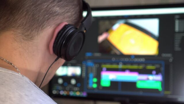 A young guy with headphones in the room is looking at a computer monitor and editing a video. He has an open program of video editing and professional color correction.