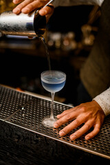 Fototapeta na wymiar bartender holds mixing cup with strainer and pours blue cocktail into glass on bar