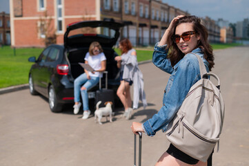 Fototapeta premium Three girlfriends travel by car with a dog and load suitcases into the trunk.