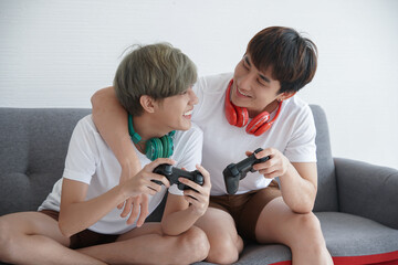 Portrait of young Asian Lgbt men couple hugging and looking at each other while they playing game together and holding joystick with earphone sitting on sofa at home