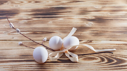 A sprig of willow with a beige ribbon, white eggs on a light brown wooden table for the Christian holiday of Easter