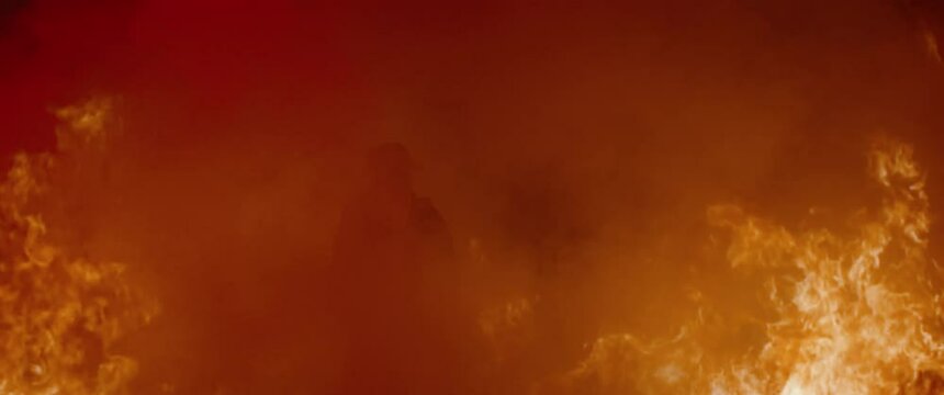 Dramatic shot of American firefighter in full gear walking into smoke and huge fire. Shot with 2x anamorphic lens 