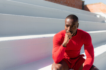 Fototapeta premium Athletic black man in sportswear sitting on stairways with joined hands looking away with serious determined face. copy space