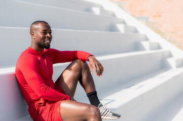 Black African athlete in sportswear resting sitting on steps with smiling happy face looking away.