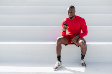 black African man with sportswear sitting on white steps using smartphone. Minimalism and copy space.