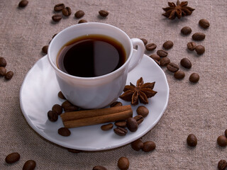 A white cup of espresso stands on a burlap table with scattered coffee beans, a cinnamon stick and stars of anise.