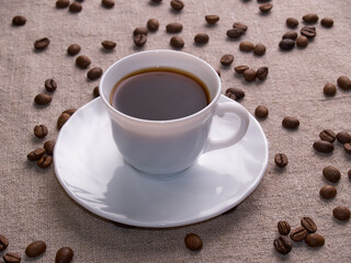 A white espresso cup stands on a burlap table with scattered coffee beans.