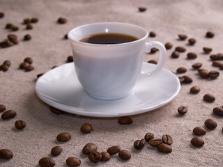 A white espresso cup stands on a burlap table with scattered coffee beans.