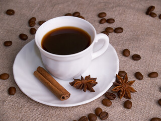A white cup of espresso stands on a burlap table with scattered coffee beans, a cinnamon stick and stars of anise.