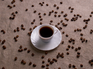 A white espresso cup stands on a burlap table with scattered coffee beans.