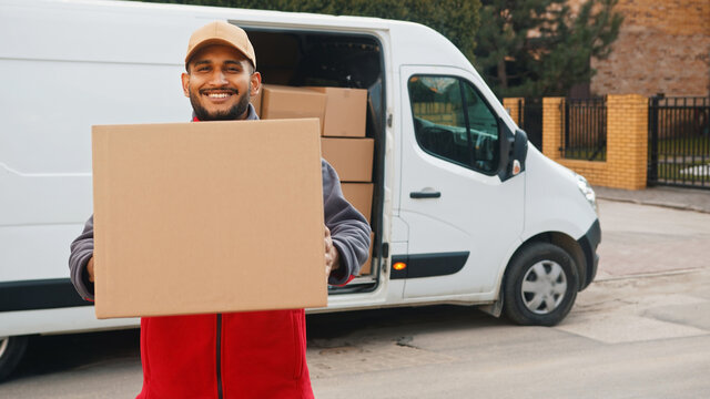 Indian man delivering package. Holding cardboard box in front of the van. High quality photo