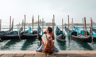 Fototapeta na wymiar Young girl traveling through Venice, sitting by the Grand Canal surrounded by gondolas with San Giorgio Maggiore in the background.