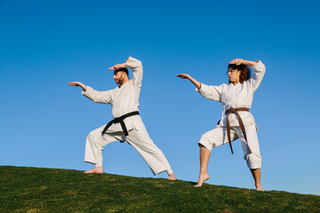 A man and a woman, dressed in karate kimonos, doing a karate stance. With a park in the background....