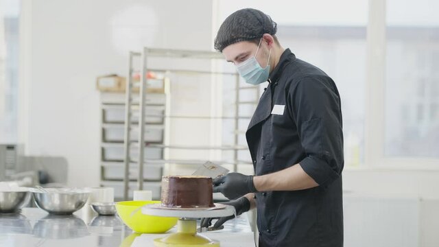 Side view of professional focused man in Covid-19 face mask spreading chocolate on round cake on rotating table. Serious concentrated Caucasian cook decorating bakery with topping in kitchen.