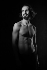 Portrait of a handsome young man with a naked torso. A man with curly hair and a beard. Black and white photo. Muscle, fitness, dance. Body sculpture. Orientation.