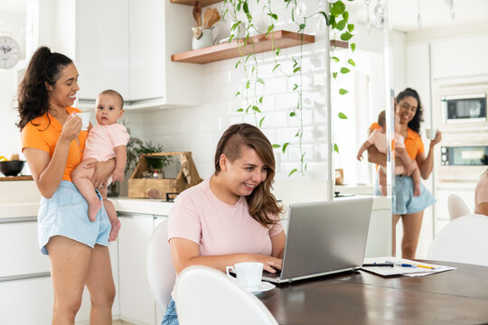 Gay Couple Working In Laptop And Other With Baby In The Kitchen.