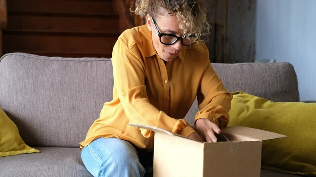 Young adult woman at home unboxing and opening a box sitting on the sofa with happiness - package delivered and discover content for pretty female people curious and excited for the article received