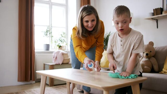 Down syndrome boy at home with mother cleaning table, helping with housework.
