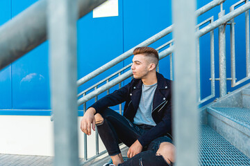 Portrait of a handsome young and rocker man sitting on stairs in the street with blue background