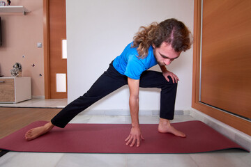 Fototapeta na wymiar Spanish bearded young male in sportswear stretching on the yoga mat at home