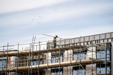 Anonymous scaffolder in PPE erecting framework of wooden planks and tall scaffolding poles high up on modern new building construction site. Professional dangerous job with safety equipment and tools.