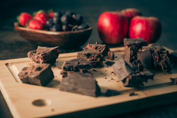 Pieces of broken assorted chocolate bar scattered on a wooden board on a wooden table. Fruits at the side. Gluten free. Gourmet. 