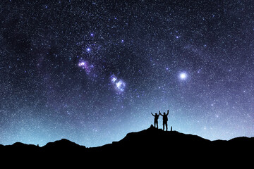 Two hikers silhouettes stands on the hill and looking at the bright starry sky orion galaxy....