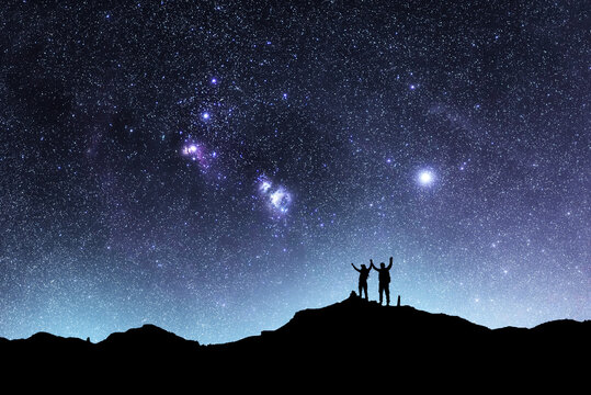 Two hikers silhouettes stands on the hill and looking at the bright starry sky orion galaxy. Beautiful fantastic night landscape.