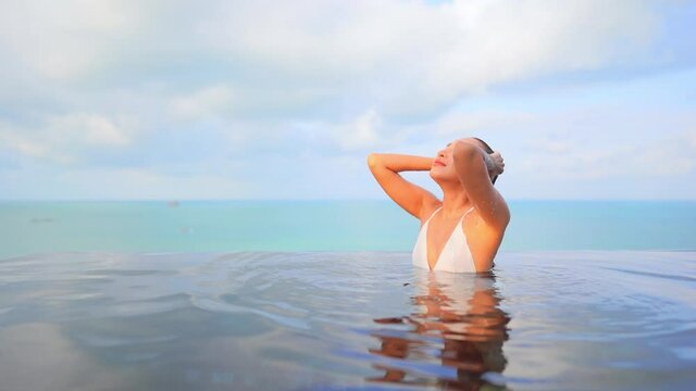 Young woman emerging from infinity pool and strokes her wet hair. Slow motion
