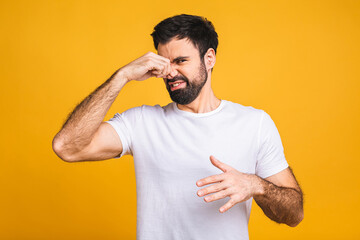 Caucasian bearded man isolated over yellow background smelling something stinky and disgusting, intolerable smell, holding breath with fingers on nose. Bad smells concept.