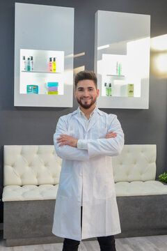 M&eacute;dico m&eacute;dico est&eacute;tico bienvenida hombre hombre brazos cruzados tienda producto blanco sonrisa sonrriente dentista barba joven guapo alegre esperando lobby consulta sala recibidor frascos pomos bata 