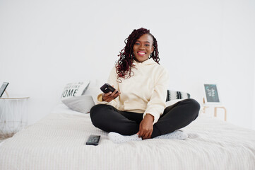 African woman watching tv at home and holding a remote control on bed.