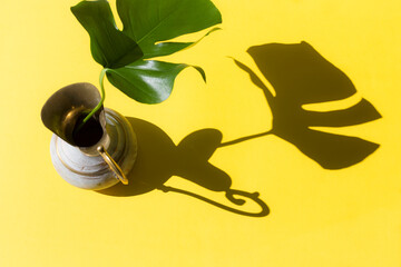 Monstera leaf in brass jug with sunlight and harsh shadow on yellow paper background. Summer concept with palm tree leaf. Copy space. Flat lay.