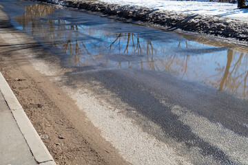 Fototapeta na wymiar Dirty asphalt road with large puddles from melting snow and a layer of sand at the sidewalk. Consequences of de-icing urban roads in winter - dust and dirt from reagents and sand mixtures