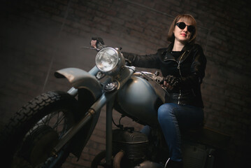 Fototapeta na wymiar Young biker woman in the sunglasses is sitting on retro motorbike in the old garage and smiles.