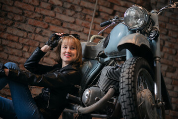 Young biker woman is sitting near the retro motorbike in the old garage and smiles.