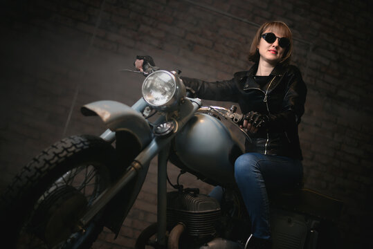 Young biker woman in the sunglasses is sitting on retro motorbike in the old garage and smiles.