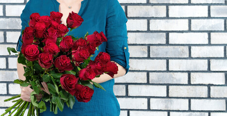 The girl received a bouquet of beautiful red roses as a gift. Flower delivery advertising banner with copy space. A girl stands against a white brick wall and holds luxurious flowers in her hands.
