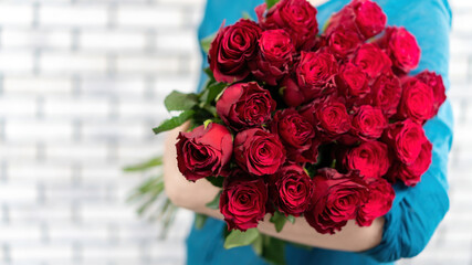 The girl is holding an armful of luxurious red roses on a white brick wall background. Greeting card with a large bouquet of red roses with copy space. Flower delivery banner.