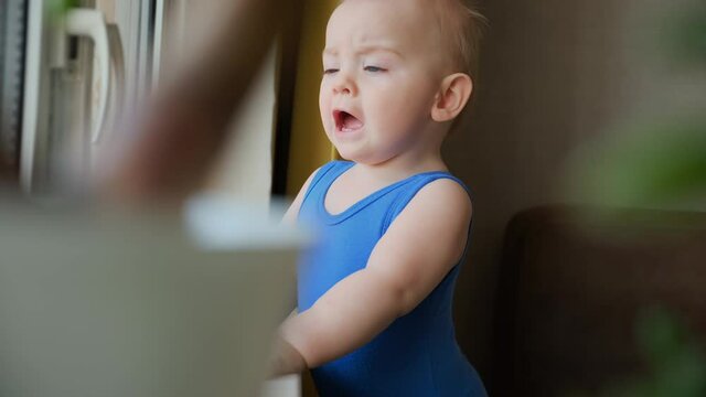 Noisy toddler boy. 11 month old serious upset baby standing on feet near window at home.