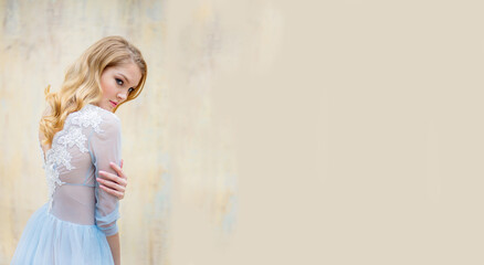 Portrait of a young blonde woman in a delicate blue wedding dress against a beige wall. She turned her back