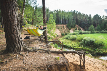 Fototapeta na wymiar A man is resting in a hammock on the high bank of the river in a pine forest and looking into the distance. A picturesque place away from the city 