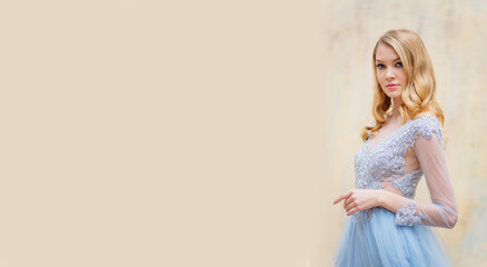 Portrait of a young blonde woman in a delicate blue wedding dress against a beige wall