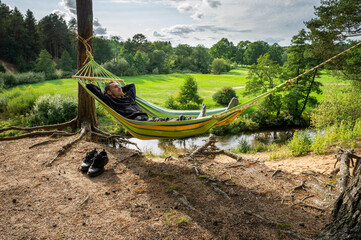 A man is resting in a hammock on the high bank of the river alone. His hiking boots stand side by...