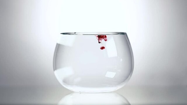 Red drops sinking in slow-motion in water in clear glass bowl with white background