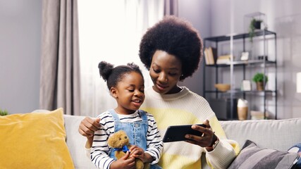 Portrait of happy African American family pretty mother and small adorable daughter sitting on sofa browsing online on smartphone watching cartoons. Mom and child searching internet on cellphone